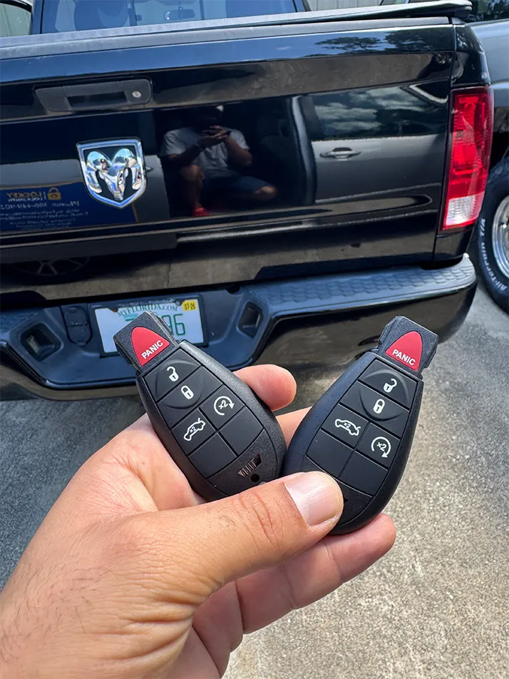 Pair of RAM FOBIK keys held behind a black RAM pickup tailgate in Palm Coast, FL